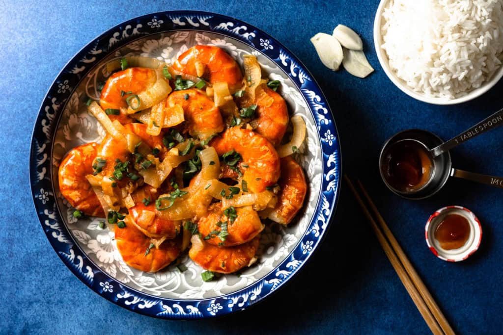Stir fried shrimp in ketchup sauce on a plate with a bowl of rice.