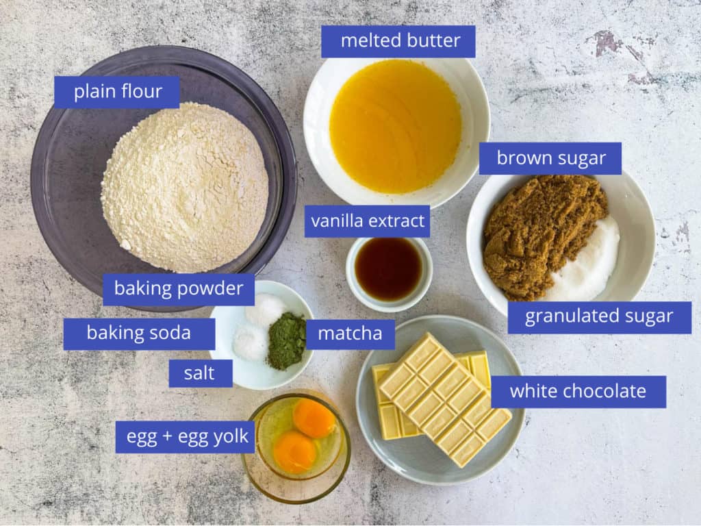 Display of ingredients for matcha cookies.  Flour, melted butter, brown sugar, granulated sugar, white chocolate, egg and egg yolk, vanilla extract, matcha powder, baking powder, baking soda, salt.